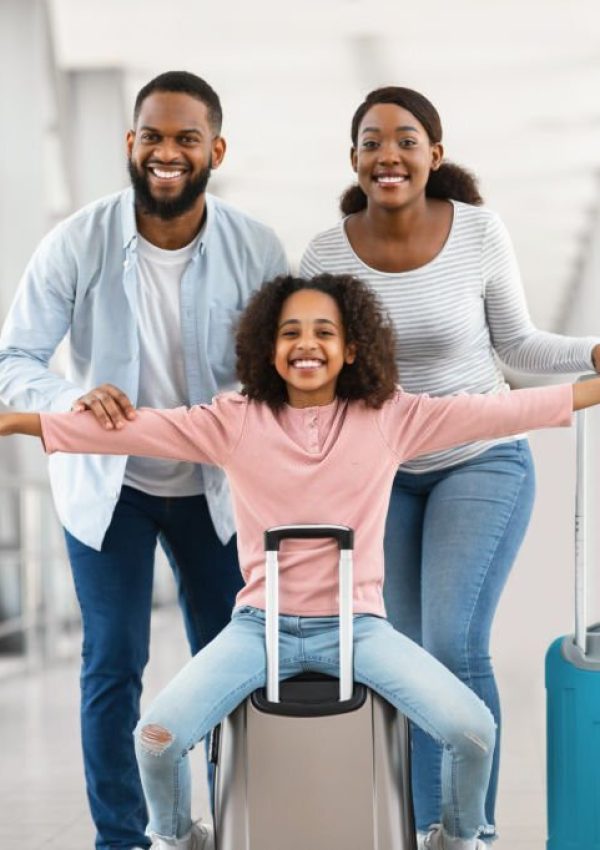 Summer Vacation Concept. Portrait of cheerful black girl having fun, sitting on suitcase and spreading hands, pretending plane flying, playing. Family of three people going on voyage, posing at camera